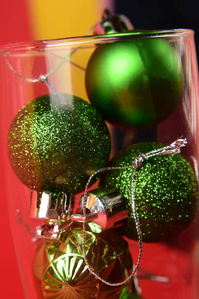 Kırmızı-sarı arka planda küçük yeşil Noel topları bir kadeh şampanya içinde yatıyor. Yakın plan.