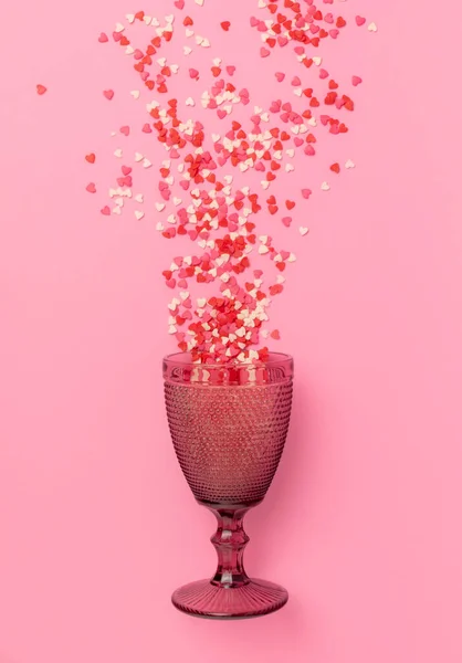 a pink glass lies on a pink pastel background. Small sweet hearts fly into the cup. The concept of bright and happy emotions. view from above