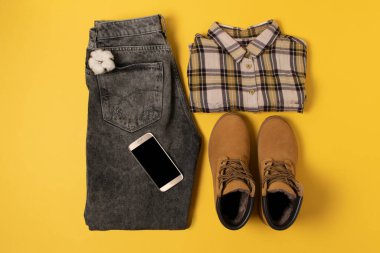 Sonbahar kıyafetlerinin ve aksesuarların sarı arka planda sıcak renklerde dizilişi. Kot, gömlek, bot ve telefon yakınlarda duruyordu.