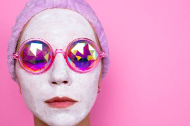 Kozmetik beyaz maskeli ve pembe gözlüklü kız pembe arka planda ciddi görünüyor. Spa konsepti