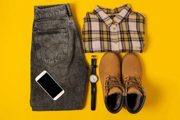 Sonbahar kıyafetlerinin ve aksesuarların sarı arka planda sıcak renklerde dizilişi. Kot, gömlek, bot, telefon ve saat yakınlarda yatıyordu.