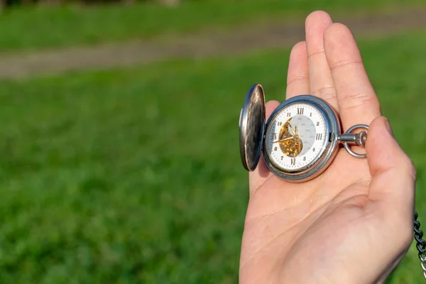 Vintage pocket watch in male hand on a background of green grass. Steampunk watch. Sunny summer day. The clock mechanism is partially visible.