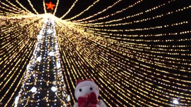 Oyuncak kardan adam, dışarıda bir sürü titreyen çelengi olan süslü Noel ağacının arka planına karşı şenlikli bir konuşma yapıyormuş gibi yakın çekim yapıyor. Mutlu Noeller ve mutlu yıllar konsepti. 4K Ultra HD