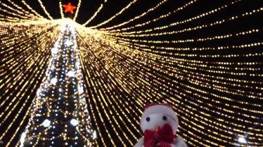 Oyuncak kardan adam, dışarıda bir sürü titreyen çelengi olan süslü Noel ağacının arka planına karşı şenlikli bir konuşma yapıyormuş gibi yakın çekim yapıyor. Mutlu Noeller ve mutlu yıllar konsepti. 4K Ultra HD