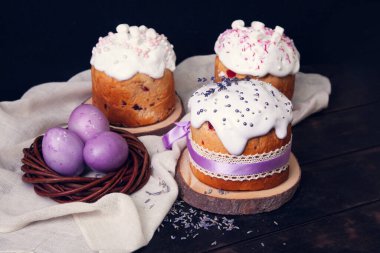 Karanlık bir arka planda paskalya pastaları, lavanta ve marşmelovla süslenmiş ve hasır bir yuvada mor Paskalya yumurtaları boyanmış..