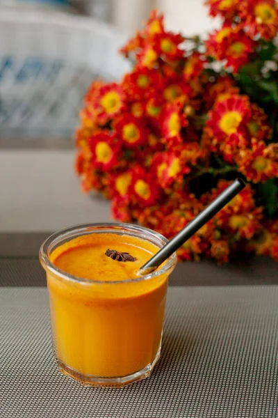 Autumn drink in glass with tube and piece of star anise on the background of bouquet of bright orange asters on the outdoor terrace. Sea buckthorn smoothie