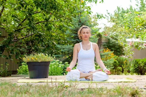 Genç bir kadın bahçede yoga yapıyor, Lotus pozisyonunda oturuyor, elleri Jana Mudra 'nın içinde katlanmış ve meditasyon yapıyor..