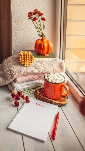 Cozy autumn still life on the windowsill with Notepad for notes: warm wool sweaters, pumpkins, maple leaves and a Cup of cocoa with marshmallows and waffles.