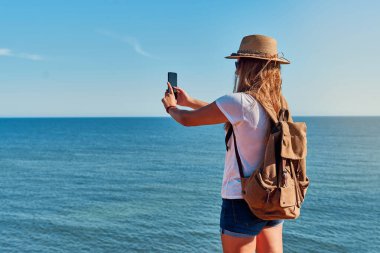 Şapkalı ve sırt çantalı genç bir kadın, yazın deniz kenarında fotoğraf çekiyor. Seyahat, tatil ve macera kavramı.