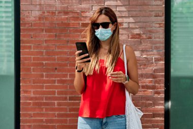 Cep telefonunu kullanan maskeli genç bir kadın. Yeni normallik kavramı ve virüse karşı savaş.