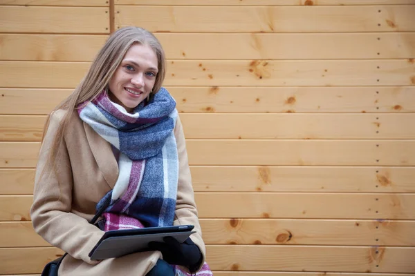 Blonde beautiful woman holding tablet outdoor. Portrait of young woman wearing tartan scarf in front of wooden wall and using tablet.