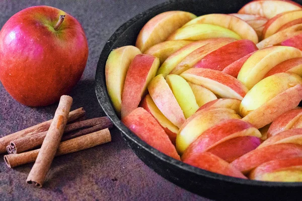 The apples cut by pieces in a pig-iron form for roasting, red apple and sticks of cinnamon on a rusty metal surface.