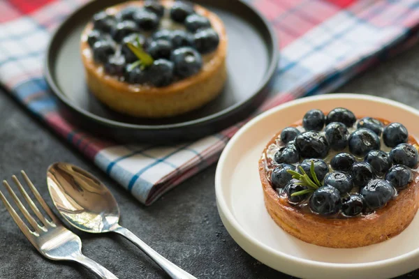 Gri bir arka plan üzerinde bir kaşık ve çatal ile bir kafes peçete siyah ve beyaz plakalar üzerinde vanilya krem ile Lezzetli yaban mersini tartalet. Üstte görüntü.