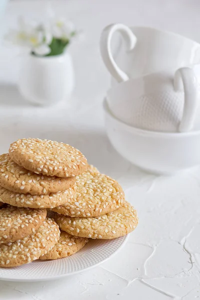 Freshly baked round sesame cookies with a apple tree flowers and three tea cups on a white table. — Stock Photo, Image