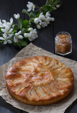 Siyah bir masada parşömen kağıdının üzerinde taze pişmiş elmalı turta. Çiçek açan bir elma ağacı dalı ve arka planda bir kutu kahverengi şeker..