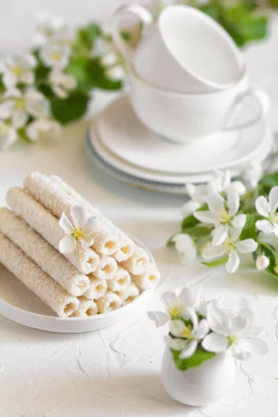 Delicati biscotti dolci sotto forma di tubi in scaglie di cocco ripieni di crema alla vaniglia su un piatto bianco e una pera gialla con due tazze da tè sullo sfondo. — Foto Stock