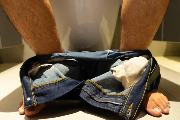 Pernas masculinas tirar jeans, sentar-se na casa de banho . — Fotografia de Stock