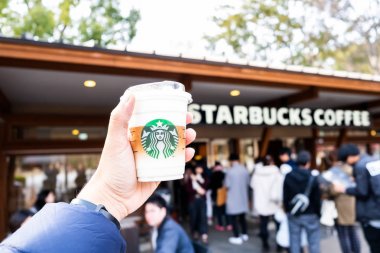 Tokyo, Japonya - 17 Mart 2019: Pembe kiraz çiçeği veya Tokyo, Japonya'nın Sakura bahar sezonunda Starbucks Coffee Ueno Onshi Park'ın önünde marka logolu Starbucks kahve fincanını tutan el.