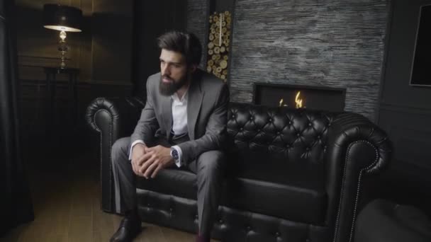暖炉のある装飾が施された壁に黒革のソファに座っているグレーのスーツでスローモーション エレガントな深刻なアゴヒゲ — ストック動画