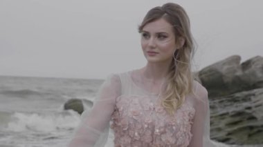 ağır çekim portre Rüzgar bulutlu gökyüzü altında güzel kız sarışın uzun saçlı kadın bakar devam kayalık deniz plaj, dalgalar