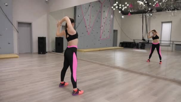 美丽的背面视图黑发女孩在运动服伸展手臂对镜子在木地板上在健身房 — 图库视频影像