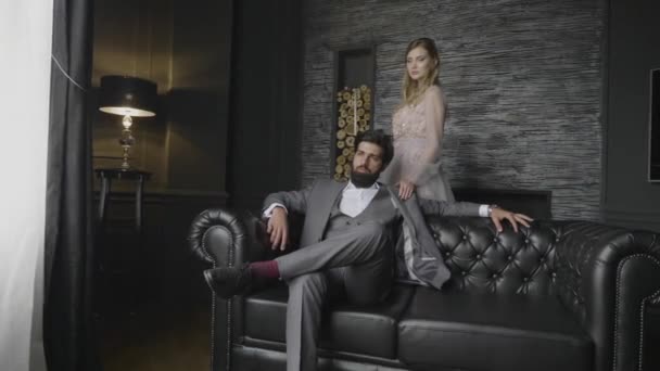慢动作严重胡须男子在灰色西装坐在皮革沙发金发女士站在后面的装饰墙 — 图库视频影像