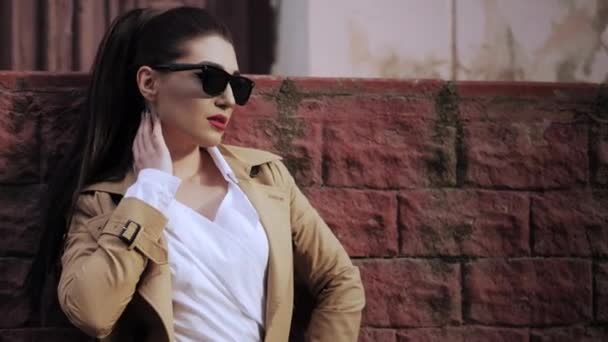Чудова леді в сонцезахисних окулярах бежеве пальто позує на цегляній стіні — стокове відео