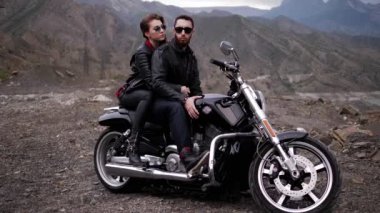 şık siyah ceketli kız yakışıklı adam motosiklet üzerinde kayalık tepe yamaçlarına karşı oturuyor