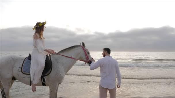 英俊男子在白色衬衫持有灰色马与长发女士吊带沿着沙滩行走 — 图库视频影像