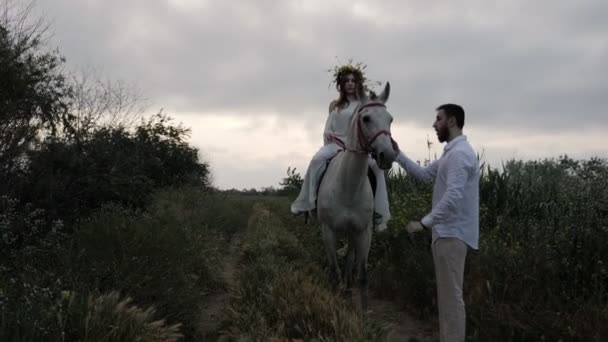 強い男は馬に座っている白い夏のドレスで女性の近くにブライドルとペットの動物によって灰色の馬を保持します — ストック動画