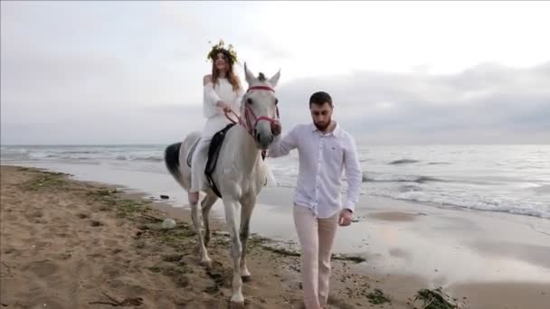 白いシャツと茶色のズボンのハンサムな男は 海の砂浜に沿って馬に乗って女性と灰色の馬をリードします — ストック動画