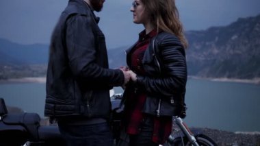 Geceleri motosiklet ve göle karşı ayakta el ile gülümseyen romantik çift