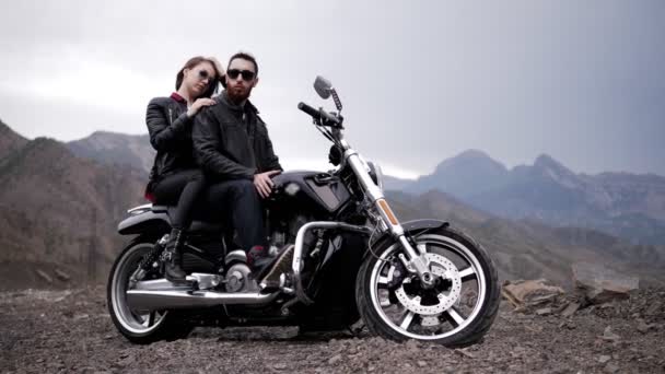 黒いドレッシングとサングラスの素晴らしいバイカーカップルは 雲の下の丘に対してオートバイに座っています — ストック動画