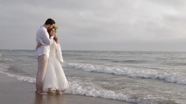 ロマンチックなカップルは 灰色の曇った空の下で海岸線に転がる泡立つ波で海水に立って抱きしめる — ストック動画