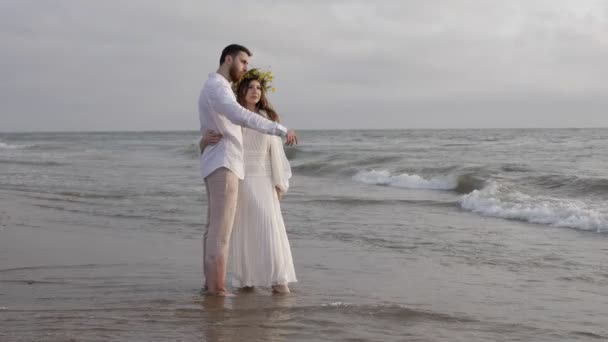 白い夏の服を着た若いロマンチックなカップルは 曇った空の下で無限の海を賞賛する水の中に立っています — ストック動画