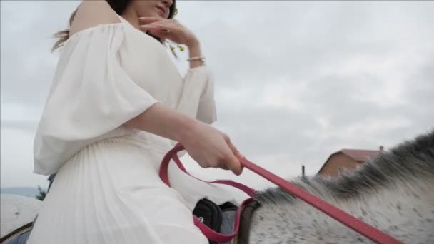 女人在长白色礼服坐在马鞍和举行吊带 英俊的男人走近马 — 图库视频影像