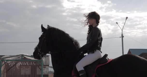 Menina bonita em jaqueta de couro senta-se no cavalo preto lento — Vídeo de Stock