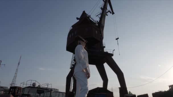 Woman poses against seaport lifting mechanism against sunset — ストック動画