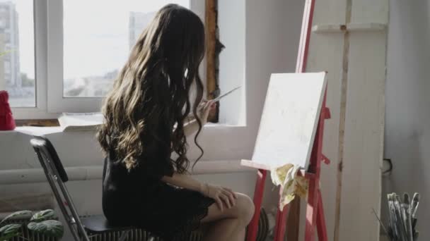 Menina em vestido preto desenha imagem com escova perto da janela — Vídeo de Stock