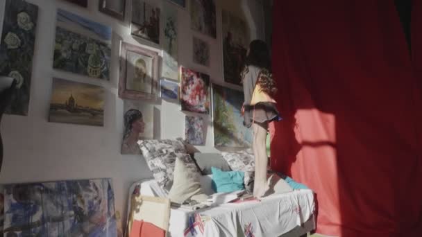 Chica consigue en sofá para mirar las pinturas en la pared en el estudio — Vídeos de Stock