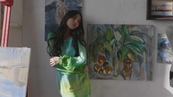 Тонка леді з довгим волоссям в зеленій сукні стоїть біля малюнків — стокове відео