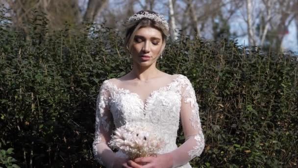 Νύφη σε φόρεμα με μπουκέτο και τιάρα κοντά σε πράσινους θάμνους — Αρχείο Βίντεο