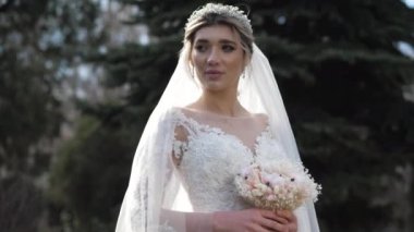 Gelin beyaz elbise giyiyor ve taç parkta buket tutuyor.