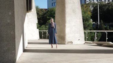 Açık renk tesettürlü ve mavi elbiseli kadın büyük sütunların yanında poz veriyor.