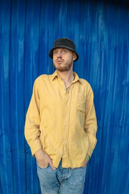 Mavi bir duvarın arkasında sarı gömlekli mutlu bir adam var..