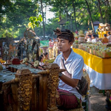 Ubud, Bali, Endonezya - 5 Mayıs 2018: Arka planda devam eden bir törenle geleneksel kıyafetler giyen geleneksel müzik aleti çalan genç bir müzisyen
