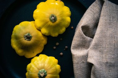 Siyah bir tavada üç sarı kabak. Dökülen biber bezelyesi. Keten havlu.