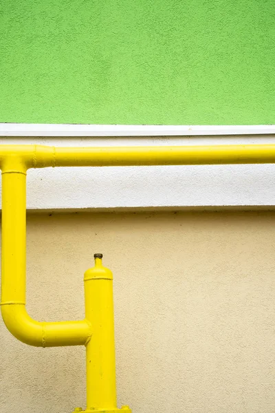 Wall of house with yellow pipe in the foreground. The wall is green and yellow with a stripe in the middle. Vertical orientation