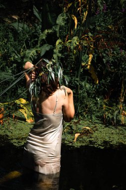 Yeşil çalılar ve ağaçlar arasında gölde ıslak ışık elbiseli kadın. Yosunlar gölde yüzer. başında söğüt dalları bir çelenk. Dikey yönlendirme