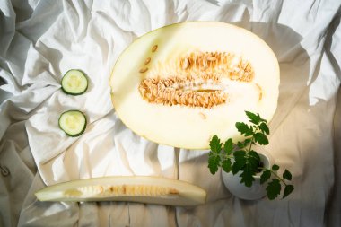 Yarım kavun, salatalık ve domates yaprakları bir vazo içinde. Kavun dilimi. Üst te. Sıcak bir ton. Kıvrımlı beyaz bir bez üzerinde. Yatay yönlendirme